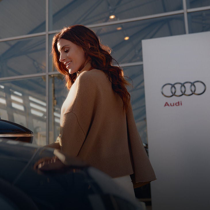 Dame in beigem Pullover steht vor einem Audi Zentrum und neben einem schwarzen Auto.